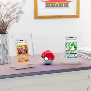 Cadre de présentation carte gradée PCA est un présentoir en acrylique transparent contenant une carte Pokémon gradée par l'entreprise PCA. On y voit un cadre transparent avec une carte Pokémon rouge de face, représentant Reptincel. Il y en adeux sur l'image sur une table. Ils sont séparés par une pokéball rouge et blanche. Sur la gauche il y a un vase avec une plante et derrière sur le mur, un cadre marron avec un dessin de maison est accroché.
