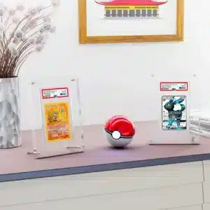 Protection acrylique pour 1 cartes PSA, résistante aux UV (99,9%) presentation est un présentoir en acrylique transparent contenant une carte Pokémon gradée par l'entreprise PSA. On y voit un cadre transparent avec une carte Pokémon rouge de face, représentant Dracaufeu. Il y en a deux sur l'image sur une table. Ils sont séparés par une pokéball rouge et blanche. Sur la gauche il y a un vase avec une plante et derrière sur le mur, un cadre marron avec un dessin de maison est accroché.