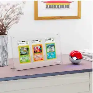 Protection cartes Pokémon PCA (99,9%) est un coffret transparent de protection pour cartes Pokémon. On y voit 3 cartes gradées par l'entreprise PCA. Le support repose sur deux petites barres rondes en métal, sur une planche avec une pokéball rouge et blanche sur la droite, devant une pile de 3 livres. Sur la gauche il y a un vase avec une plante et derrière sur le mur, un cadre marron avec un dessin de maison est accroché.
