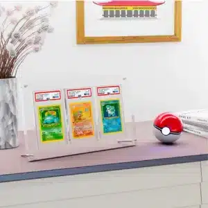 Protection acrylique pour 3 cartes PSA, résistante aux UV (99,9%) est un coffret transparent de protection pour cartes Pokémon. On y voit 3 cartes gradées par l'entreprise PSA. Le support repose sur deux petites barres rondes en métal, sur une planche avec une pokéball rouge et blanche sur la droite, devant une pile de 3 livres. Sur la gauche il y a un vase avec une plante et derrière sur le mur, un cadre marron avec un dessin de maison est accroché.