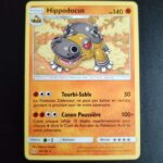 Hippodocus 69/156. Carte du TCG Pokémon issue de la série Ultra-Prisme du bloc Soleil et Lune (SL05, SL: Ultra-Prisme), parue le 02/02/2018. Carte Pokémon de Niveau 1 de type Combat, évolution de Hippopotas. Rare. Illustrateur: Shigenori Negishi. L'illustration représente hippopotame avec des parties du corps métalliques, notamment son dos d'où sortent des jets de liquide jaunâtre via des orifices circulaires.
